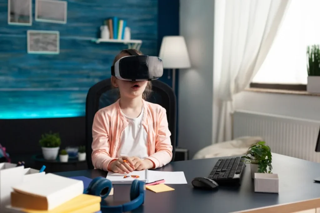 A young girl wearing a virtual reality headset sits at a desk with papers, crayons, and a computer keyboard in a modern home office.