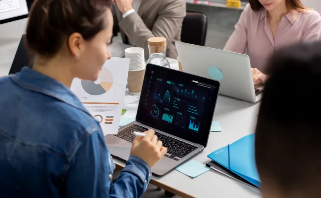 A woman in a business meeting, using a laptop displaying colourful data analytics, seated amongst colleagues in an office environment.