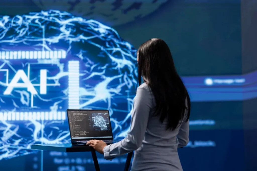 A woman in a white blouse is standing while using a laptop, facing a large screen displaying a futuristic neural network graphic in a high-tech environment.