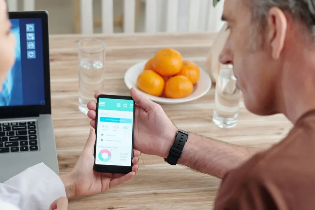 A man is checking a health app on his smartphone, which displays various fitness metrics. He is seated at a wooden dining table where a plate of oranges and glasses of water are also visible.